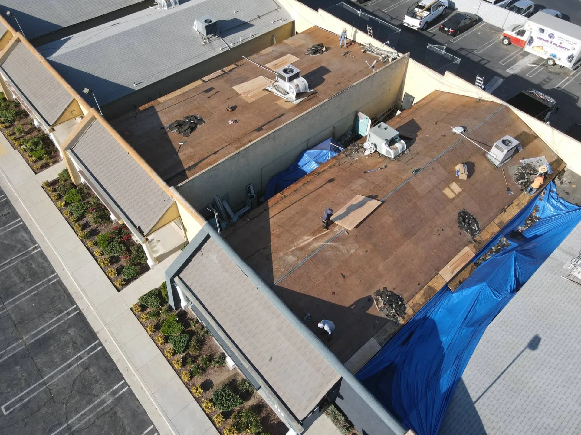 Aerial view of a retail strip center during a flat roof replacement: roof stripped to the plywood deck, rooftop HVAC units, tarps along the edges and roofers at work