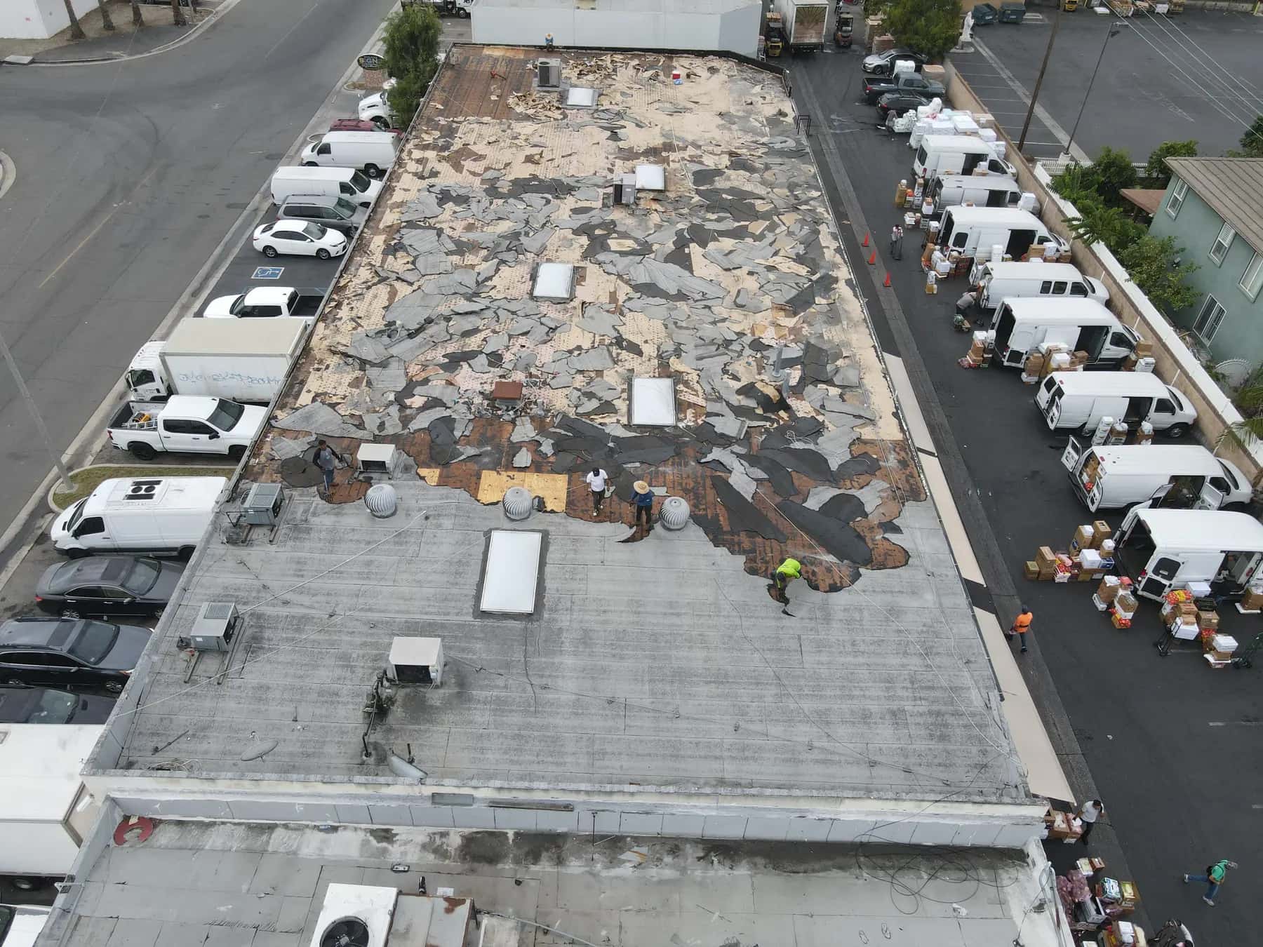 Roofers tearing off the last section of the old gray cap sheet by the turbine vents, with the loading yard busy below