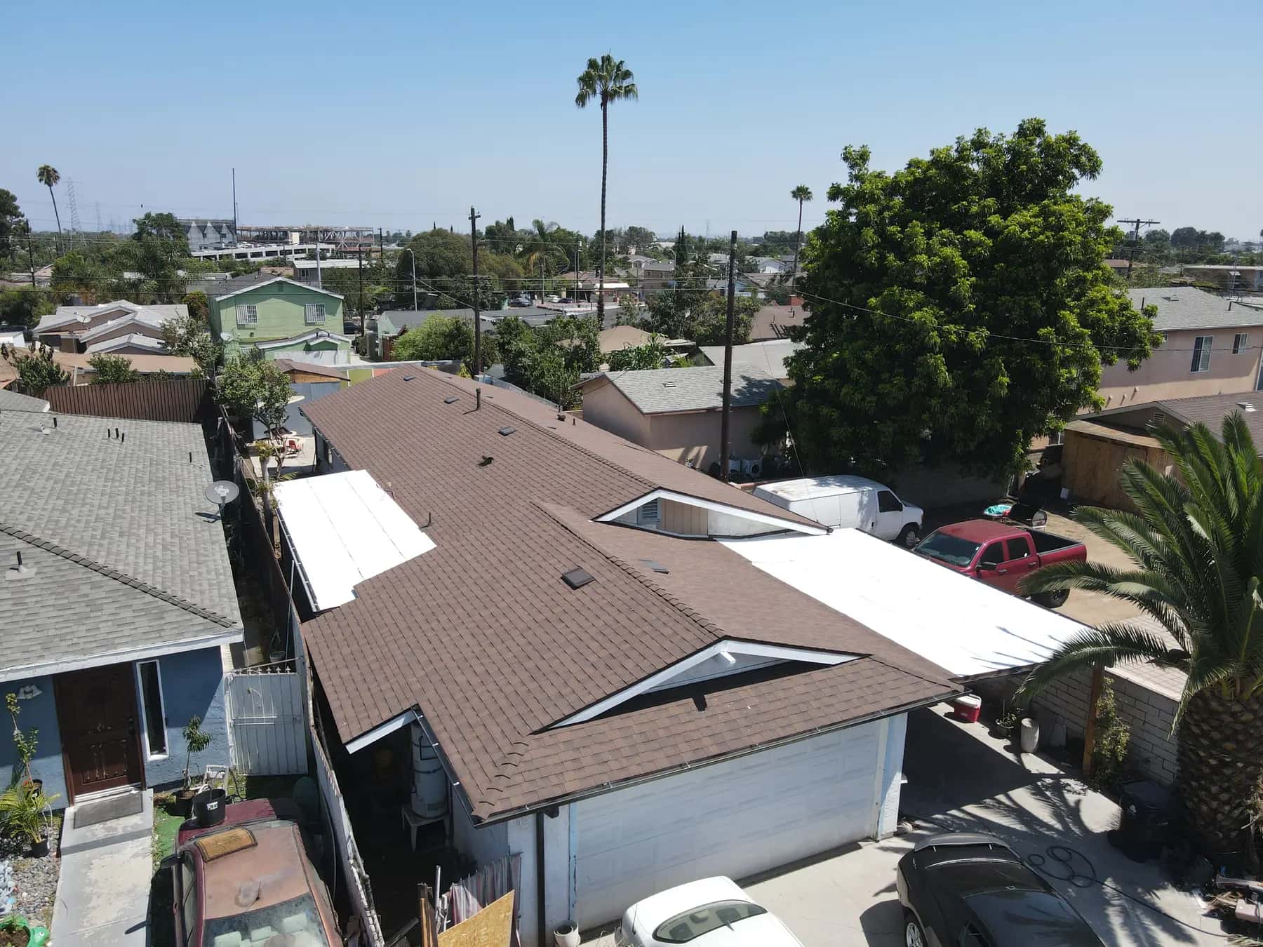 Aerial view of a long single-story home in South Los Angeles with a new brown shingle roof, white flat roof sections at the sides and a palm tree beside it