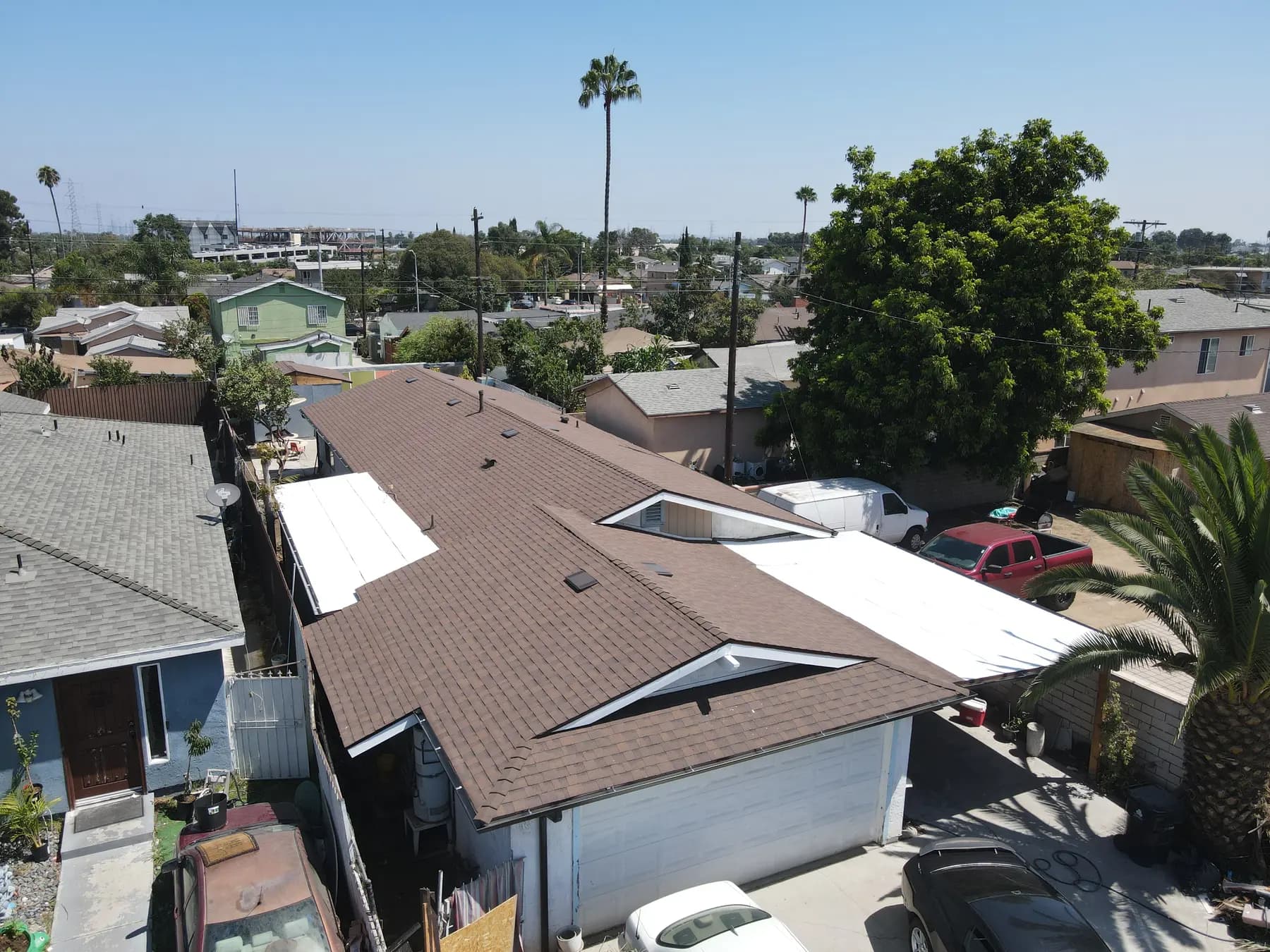 Aerial view of a long single-story home in South Los Angeles with a new brown shingle roof, white flat roof sections at the sides and a palm tree beside it