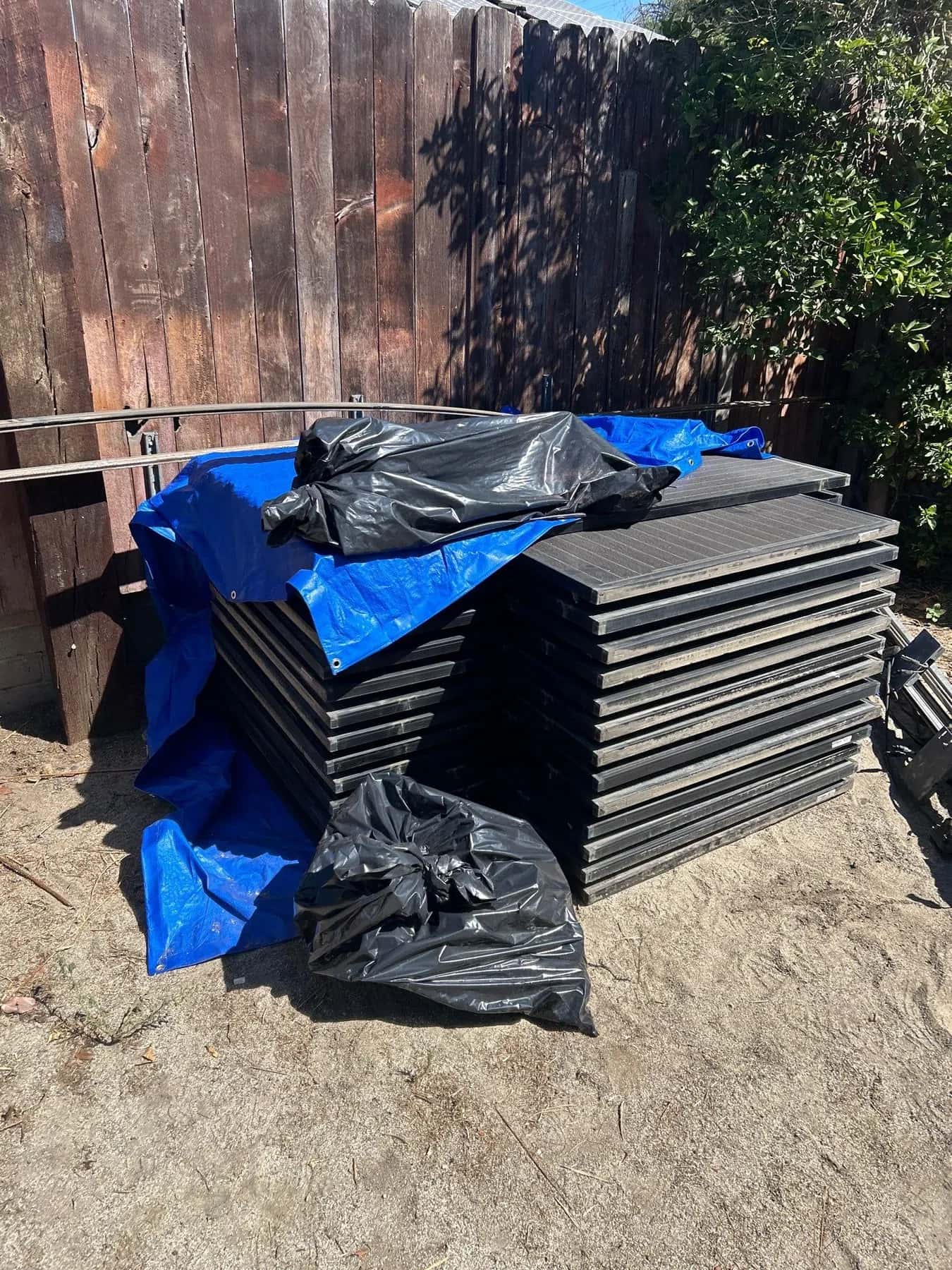The solar panels stacked flat on boards in the yard, under a blue tarp and kept clear of the work