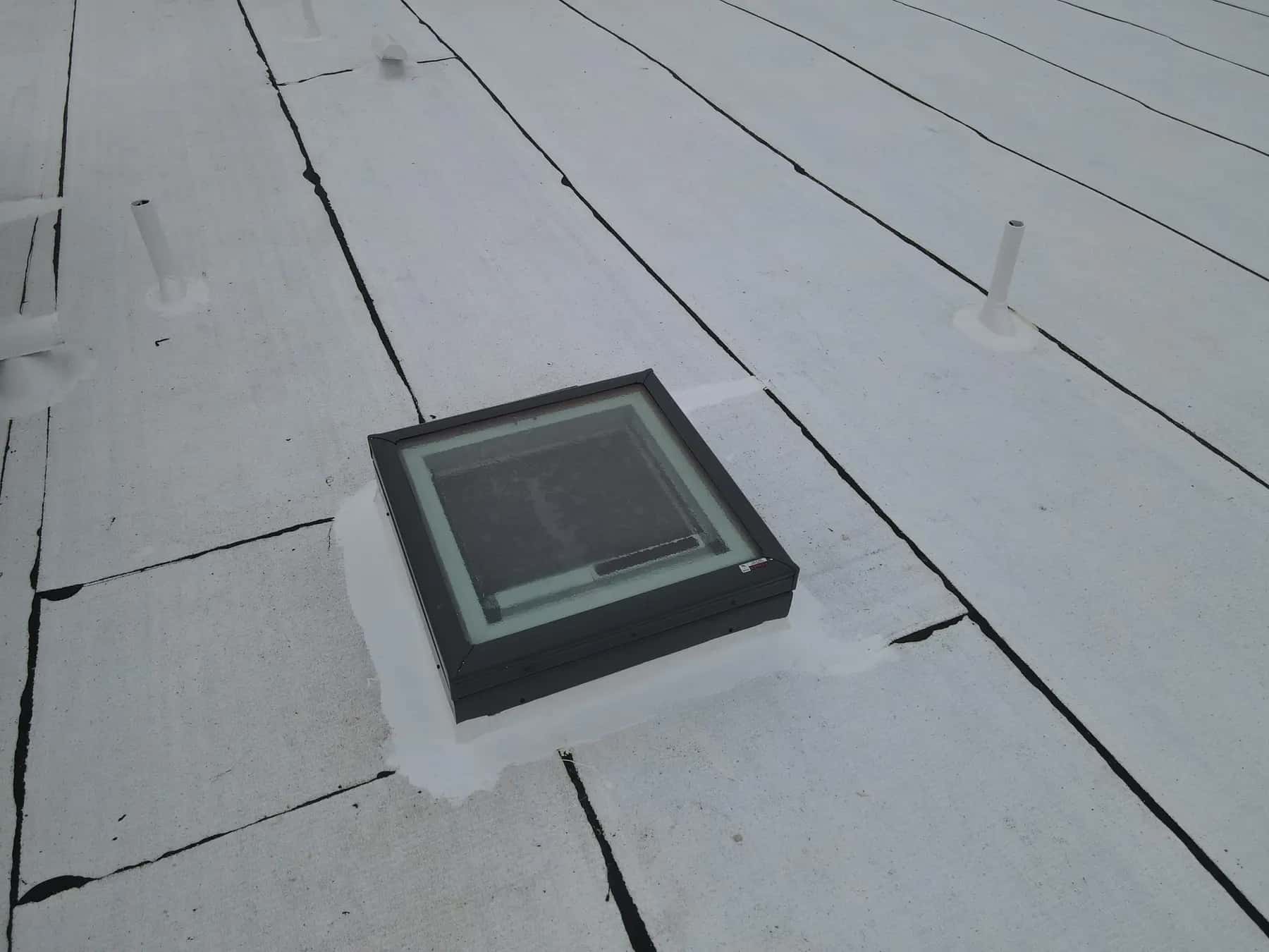 Skylight on a white-finished curb, with plumbing vents and dark seam lines running between the roof sheets
